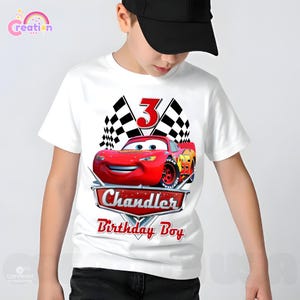 May include: White t-shirt featuring a red race car graphic with the number 3, checkered flags, and the text "Chandler Birthday Boy." The design includes the Lightning McQueen character from the Cars movie.