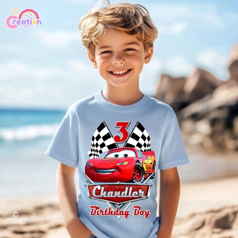 May include: Light blue t-shirt featuring a red race car graphic, a checkered flag, and the number 3. The shirt also includes the name "Chandler" and the text "Birthday Boy".