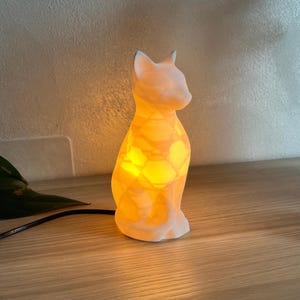 Puede incluir: Lámpara con forma de gato que emite una luz amarilla cálida. La lámpara blanca tiene un patrón texturizado y está colocada sobre una superficie de madera. Está enchufada con un cable negro.