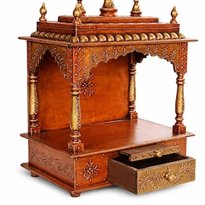 May include: A wooden prayer shrine with detailed carvings and a warm brown finish. The shrine has a small drawer, ornate pillars, and a decorative top with gold accents. The design is detailed with floral patterns.