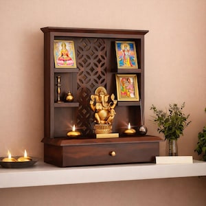 Puede incluir: Un altar de madera marrón oscuro con un cajón, estantes y un recorte decorativo. Presenta imágenes religiosas enmarcadas, una estatua dorada de Ganesha, velas y pequeños objetos decorativos. El altar está sobre una estantería blanca.