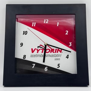 May include: Square black framed clock with a white face. The clock face has a red and white diagonal design with the word "VYTORIN" and the text "(ezetimibe simvastatin) tablets". The clock hands are black with a gold center.
