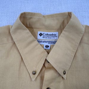 May include: Close-up of a tan button-down shirt with a collar. The shirt has a label that reads "Columbia Sportswear Company, Portland, Oregon, USA." The label also indicates the shirt is XL and "MEN'S."