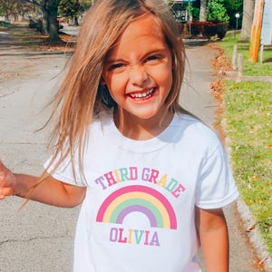 May include: A young girl wearing a white t-shirt with a rainbow design and the words "THIRD GRADE OLIVIA" printed on it. She is smiling and pointing to the left. She is also wearing pink gingham shorts.