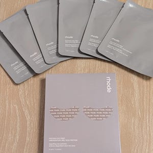 Peut inclure: Un ensemble de patchs cosmétiques gris pour les yeux et une boîte assortie. La boîte et les paquets individuels sont étiquetés "rhode" et "PEPTIDE EYE PREP". L'emballage est de couleur gris clair.