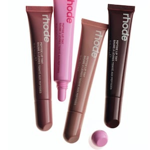 Op de afbeelding: Vier tubes rhode peptide lip tint in verschillende tinten roze, mauve en diepe bessenkleuren. Elke tube toont de merknaam, productnaam en een volume van 10 ml op de voorzijde. Eén roze dop is losgemaakt en ligt onderaan de tubes afgebeeld.