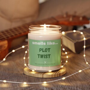 May include: A lit candle in a glass jar with a green label that reads "smells like... PLOT TWIST". The candle sits on a round bamboo coaster, surrounded by string lights. A guitar and a book are in the background.