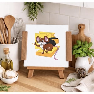 May include: A cartoon illustration of a brown mouse with a black cape holding a small object, standing on a yellow cheese block. The artwork is displayed on a white canvas, supported by a small wooden easel. The background includes kitchen utensils and decor.