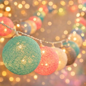 Op de afbeelding: Een slinger van kleurrijke, ronde decoratieve lampjes. De lampjes zijn in verschillende kleuren, waaronder teal, roze en geel. Elk lampje is gemaakt van geweven materiaal en geeft een warme gloed. De achtergrond is vervaagd met bokeh-lichtjes.