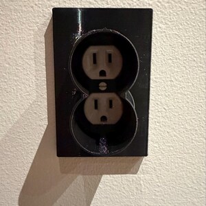 May include: A black electrical outlet with two receptacles is mounted on a textured white wall. The outlet has a rectangular faceplate and a shadow cast on the wall. The receptacles are light beige.