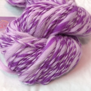 May include: A close-up of a skein of yarn in shades of purple and white. The yarn is thick and textured, with a soft, fluffy appearance. The colors are swirled together, creating a marbled effect. The yarn is likely for knitting or crocheting.