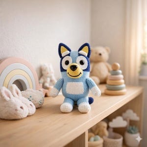 Puede incluir: Muñeco Bluey tejido a ganchillo, cuerpo azul claro con detalles blancos en el pecho, patas y pies. La cabeza es azul oscuro con orejas amarillas y una cara sonriente. En una estantería de madera con otros juguetes.