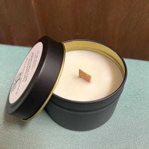 May include: A black candle tin with a gold rim and a wooden wick. The lid is partially open, revealing the creamy white candle wax. The label on the lid includes text and a botanical illustration. The candle sits on a light blue surface.