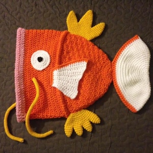 May include: An orange crochet fish-shaped hat with white and yellow accents. The hat features a white eye, a white patch, and yellow fins. The hat is designed with a pink trim and yellow tendrils.