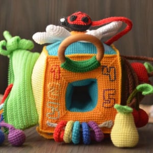 Può includere: Un giocattolo per bambini fatto a mano all'uncinetto dai colori vivaci. Il giocattolo è un cubo con un anello di legno, una coccinella e vari elementi all'uncinetto attaccati, tra cui una foglia verde, una pera e anelli colorati. Il cubo ha i numeri 1, 4 e 5.