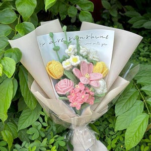 May include: A crocheted bouquet of flowers in shades of pink, yellow, and white, wrapped in tan paper. The bouquet includes tulips, roses, and other floral designs. A card with the text "You are sunshine, love with you" is visible.