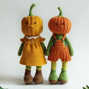 May include: Two handmade crochet dolls with pumpkin heads. One doll wears a yellow dress with a white collar and brown boots. The other doll wears orange overalls and green shoes. Both dolls have green stems.