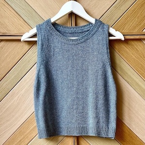 Puede incluir: Un chaleco sin mangas de color azul grisáceo con cuello redondo, colgado de una percha de madera blanca. El chaleco es de punto. El fondo es una puerta de madera con un patrón geométrico.