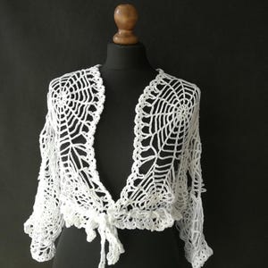 May include: A white, open-front crochet shrug with a spiderweb design. The bolero-style garment features a delicate, lacy pattern and is tied at the front. The sleeves are three-quarter length, and the shrug is displayed on a black mannequin.