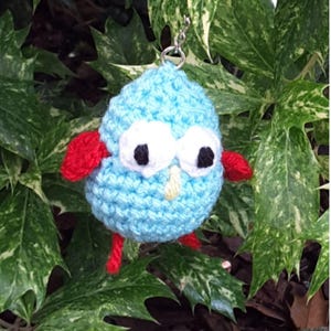 May include: A handmade blue crochet bird keychain with large white eyes, black pupils, and red wings and feet. The bird has a yellow beak and is hanging from a silver keychain. The background is green foliage.
