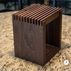 May include: A dark brown wooden knife block with a rectangular frame and a series of vertical slots along the top edge. The structure features a visible wood grain pattern and an open side design. A small circular logo with the text Furniast is visible.