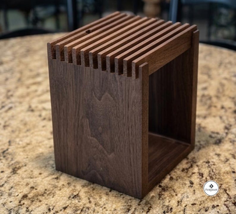 May include: A dark brown wooden knife block with a rectangular frame and a series of vertical slots along the top edge. The structure features a visible wood grain pattern and an open side design. A small circular logo with the text Furniast is visible.