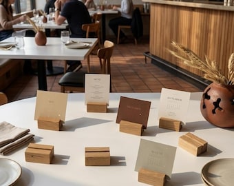 May include: A collection of small rectangular wooden card holders arranged on a white table. Several holders contain printed calendar cards for months like May, September, and August, while others remain empty. A menu card is also displayed.