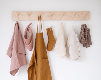 May include: A light-colored wooden wall rack with seven pegs holds a collection of items: a dusty rose cloth, a matching knotted bandana, a mustard yellow apron, a single ribbed sock, a cream-colored knit cloth, a button-up cardigan, and gloves.