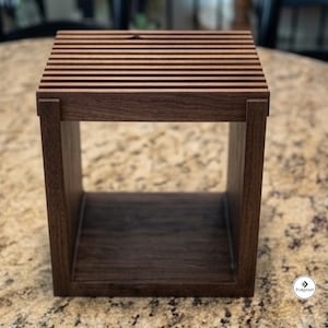 May include: A square, dark brown wooden stand featuring a slatted top surface with horizontal bars and a hollow, open-front base. The structure is constructed with visible joinery at the corners, showcasing a natural wood grain texture throughout.