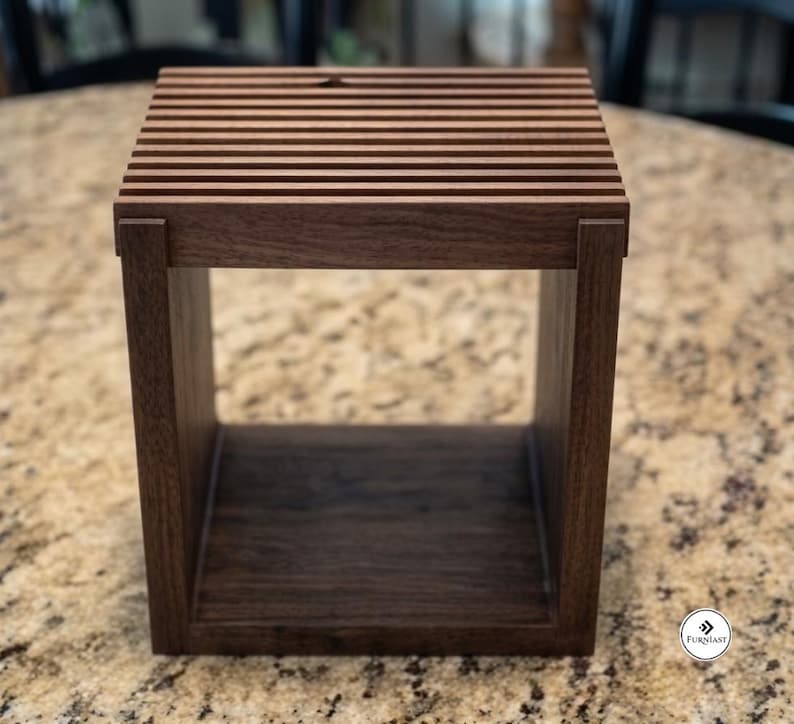 May include: A square, dark brown wooden stand featuring a slatted top surface with horizontal bars and a hollow, open-front base. The structure is constructed with visible joinery at the corners, showcasing a natural wood grain texture throughout.