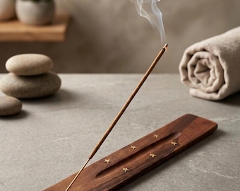 May include: A rectangular dark brown wooden incense holder featuring a carved groove and small inlaid gold-colored star accents. A single lit incense stick is inserted into the holder, emitting a thin trail of white smoke into the air.