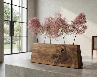 May include: A rectangular wooden vase with a natural grain pattern and a visible knot, featuring four holes along the top edge. Four stems of fluffy, light pink smoke bush flowers are inserted into the holes, standing upright in the wooden block.