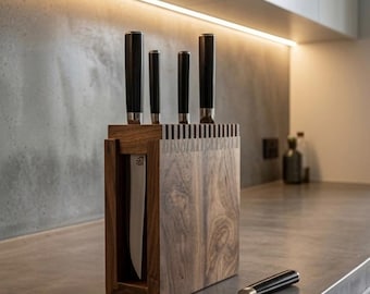 May include: A rectangular dark wood knife block with a visible grain pattern, featuring vertical slots holding four knives with black handles. One additional knife with a matching black handle lies horizontally on the surface in front of the block.