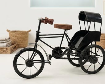 May include: A black metal tricycle model featuring a brown wooden seat and brown handlebar grips. The vehicle includes a rear passenger canopy, thin spoked wheels, a chain drive mechanism, and a frame structure resembling a traditional rickshaw.