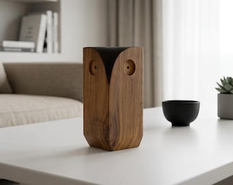 May include: A wooden owl figurine with a natural brown wood grain body and a smooth black top section. The owl features two circular recessed eye sockets carved into the front face, creating a stylized representation of an owl in a vertical form.