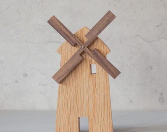 May include: Wooden windmill figurine with a light brown body and dark brown blades. The miniature windmill has a small window and door cut out of the front. The blades are attached with a small, round, brown piece.