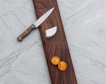 May include: A long, rectangular dark brown walnut wood serving board with a handle cutout at the top. A silver knife with a wooden handle rests on the board next to a wedge of white cheese and two slices of orange cherry tomatoes.
