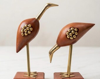 May include: Two bird figurines crafted from polished brass and smooth brown wood. Each bird features a wooden body with a cluster of small brass spheres, a brass neck and head, and thin brass legs standing on a square wooden base.