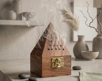 May include: A pyramid-shaped wooden incense burner with a dark brown finish and a brass-colored metal latch on the front. The top section features small, teardrop-shaped cutouts that allow thin wisps of white smoke to rise from the structure.