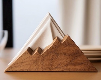 May include: A triangular wooden napkin holder with a natural wood grain finish, featuring a stepped mountain-like silhouette. A folded white paper napkin is inserted into the center slot, mimicking the appearance of a snow-capped mountain peak.