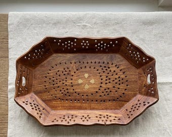 May include: An octagonal wooden tray featuring intricate laser-cut patterns along the sides and base. The tray is crafted from a medium-brown wood with a visible grain and includes a central brass inlay shaped like a four-petaled flower.