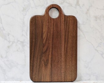 May include: A rectangular dark brown wooden cutting board with a smooth finish and a circular handle cutout at the top center. The board features a visible natural wood grain pattern throughout its surface and has rounded corners at the bottom.