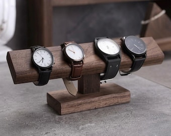 May include: A dark brown wooden watch display stand with a horizontal bar supported by a central pillar and a rectangular base. Four wristwatches with various colored faces and leather or metal straps are wrapped around the horizontal bar.
