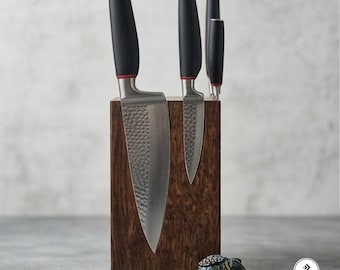May include: A set of four kitchen knives with hammered metal blades and black handles with red accents, held by a rectangular dark wood magnetic knife block. A small, colorful, patterned decorative figurine sits in front of the wooden block.