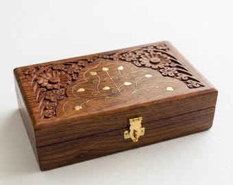 May include: A rectangular dark brown wooden box featuring intricate floral carvings on the lid and a central inlaid brass vine pattern. The box includes a polished gold-tone metal latch on the front, securing the lid to the base of the container.