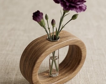 May include: An oval-shaped wooden vase with a natural grain finish featuring a central clear glass test tube insert. The vase holds three dark purple carnation stems, including one fully bloomed flower and two small closed buds.