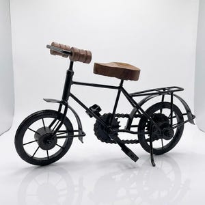May include: A decorative miniature bicycle model constructed from black metal with a dark brown wooden seat and matching brown wooden handlebar grips. The bicycle features a detailed chain, spoked wheels, a rear rack, and a kickstand for stability.