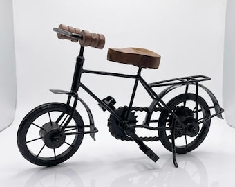 May include: A decorative miniature bicycle model constructed from black metal with a dark brown wooden seat and matching brown wooden handlebar grips. The bicycle features a detailed chain, spoked wheels, a rear rack, and a kickstand for stability.