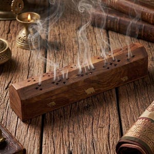 May include: A rectangular wooden incense burner box with a dark brown grain, featuring brass elephant inlays on the side and star-shaped cutouts on the lid, with thin wisps of white smoke rising from the top openings of the wooden container.