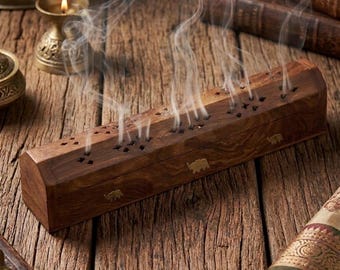 May include: A rectangular wooden incense burner box with a dark brown grain, featuring brass elephant inlays on the side and star-shaped cutouts on the lid, with thin wisps of white smoke rising from the top openings of the wooden container.